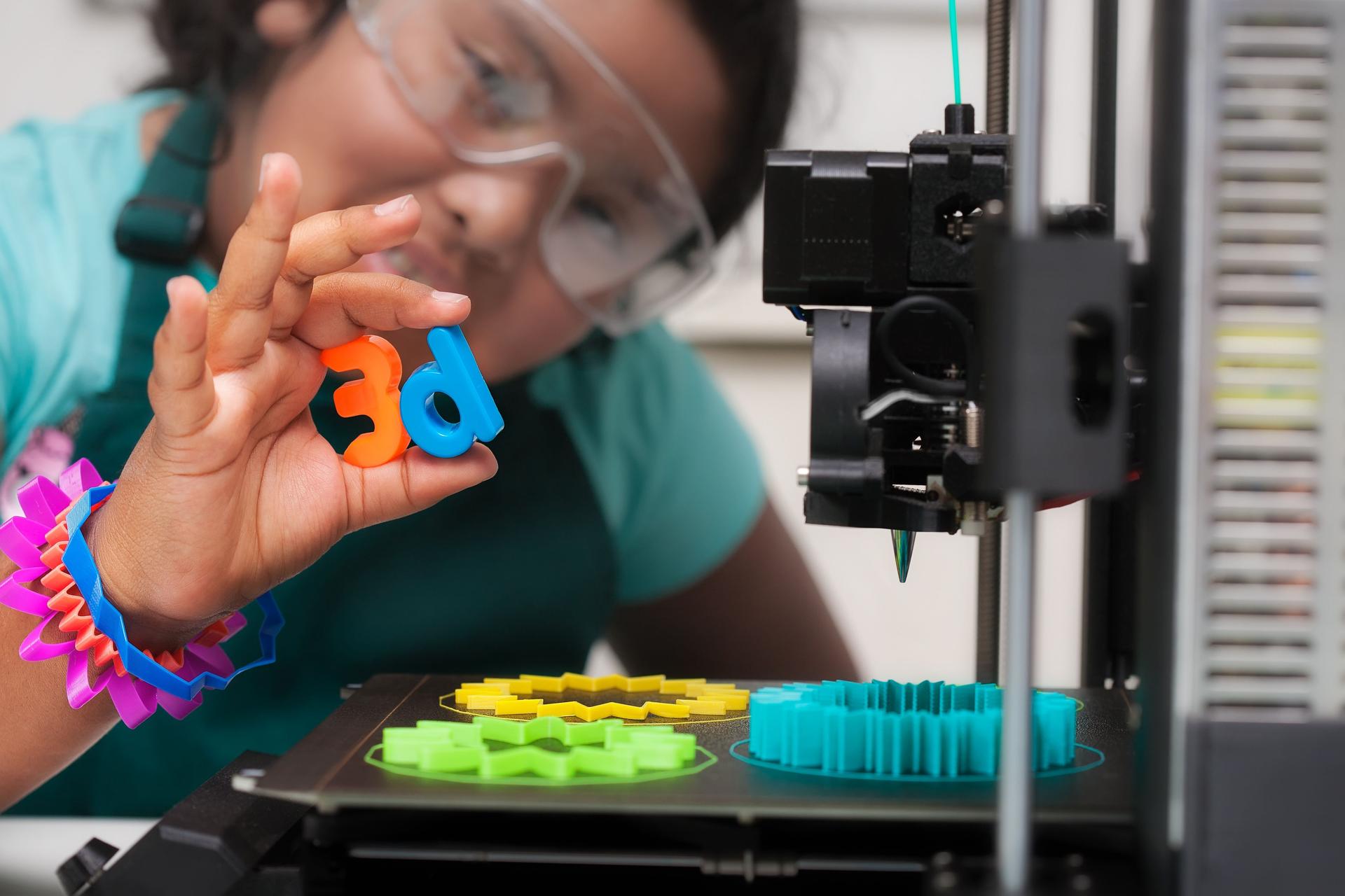 A smart latino student wearing multi-colored 3d printed shapes as jewelry, next to a 3d printer with designs on the heated print bed, and holding 3d letters in her hand.